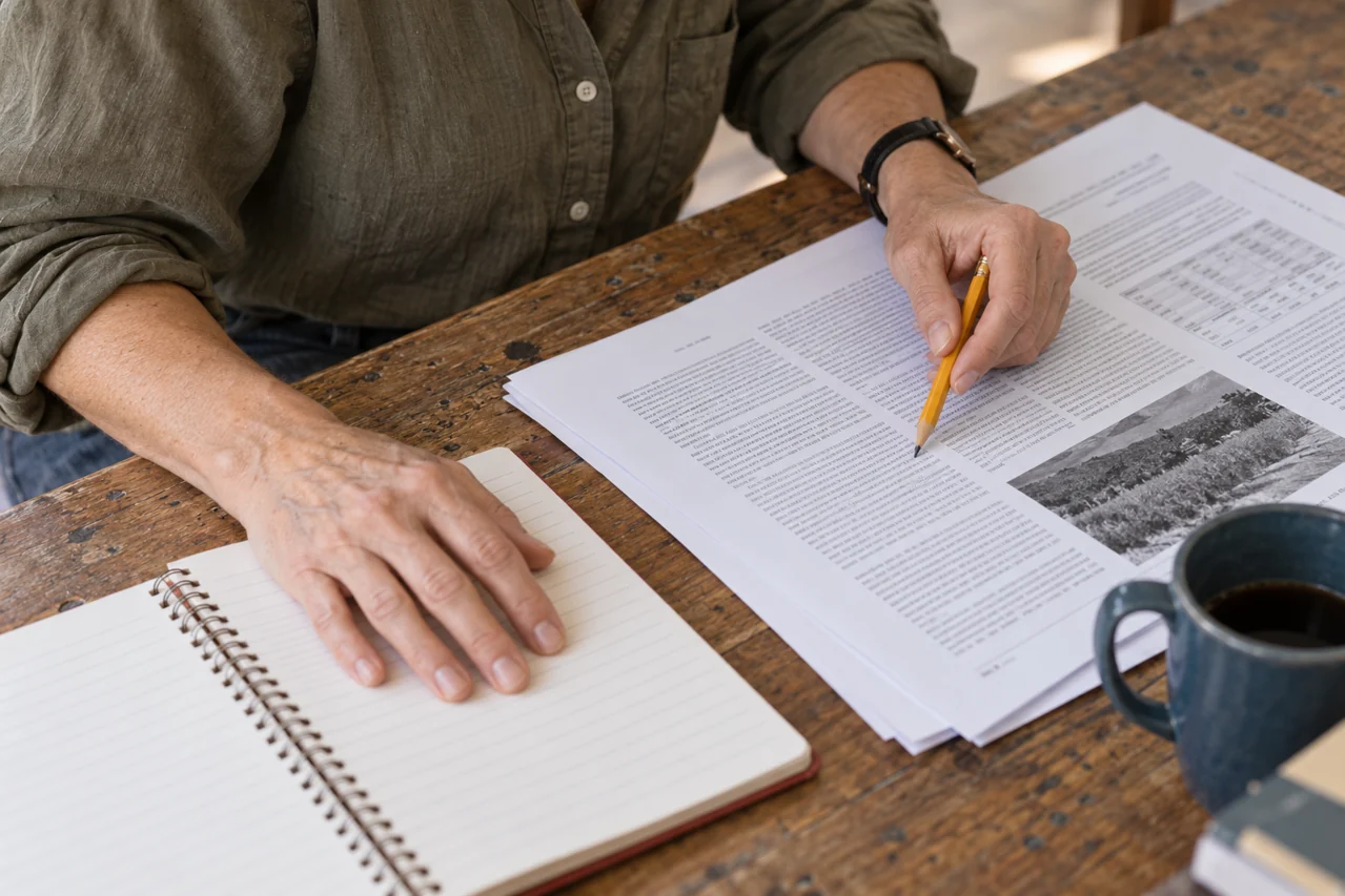 Close view of hands, a pencil, a research paper and an open lined notebook on a worn oak table.